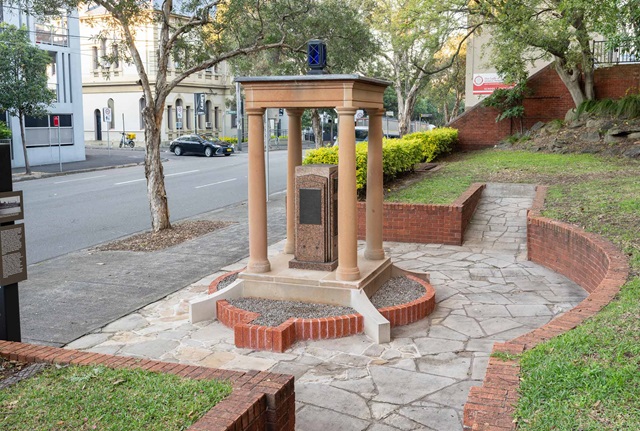 A red granite pillar in an open sandstone structure made of four columns and a roof. The structure is in a paved area surrounded by a low red brick wall in a park, next to a city street.