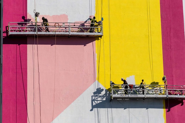 Workers on suspended platforms paint a large building facade with pink and yellow geometric patterns.