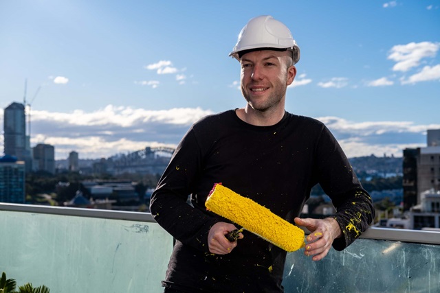 A man with hard hat, holding a paint roller coloured yellow, stands on a balcony against a city skyline with blue sky.