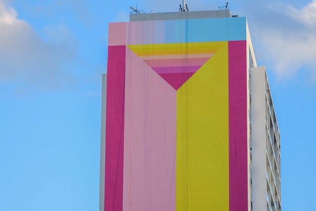 A tall building features a colorful geometric mural with pink, yellow, and blue triangles against a clear blue sky.