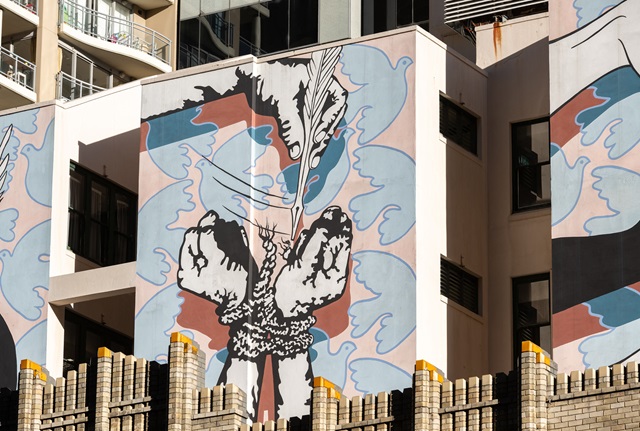 A mural on the outside wall of a building depicting 2 hands bound together with rope being freed by a third hand holding a quill pen. The background of the mural is a field of flying blue doves.