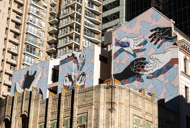 Three walls on a building are painted with murals depicting motifs of peace. The mural is several stories high, and in front of the painted building is another ornate brick building. In the background are many skycrapers.
