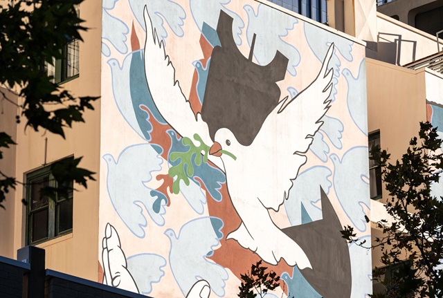 A mural on the outside wall of a building depicting a dove holding an olive branch in its beak in front of a falling bomb.