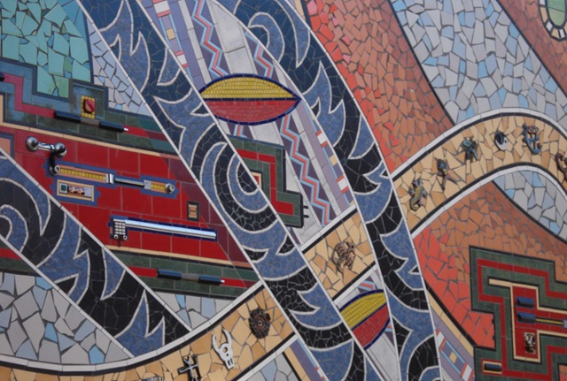 A large and colourful mosaic depicting geometric shapes and patterns made from ceramic tiles, with three-dimensional ceramic pieces amongst the tiles.