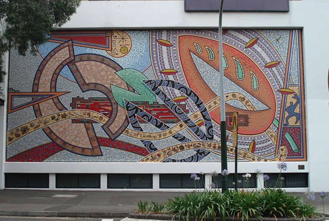 A large mosaic depicting geometric shapes and patterns made from ceramic tiles, on a wall next to a footpath and small flowerbed.