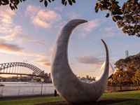 A fish hook shaped sculpture on the grass with the harbour bridge in the background.