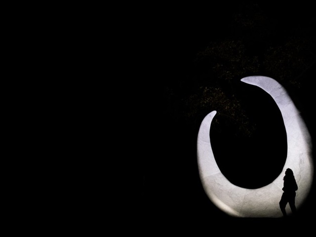 A fish hook shaped sculpture on a black background, with the silhouette of a person walking in front of it.