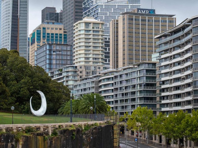 A fish hook shaped sculpture on a lawn in a park, with many city skyscrapers in the background.
