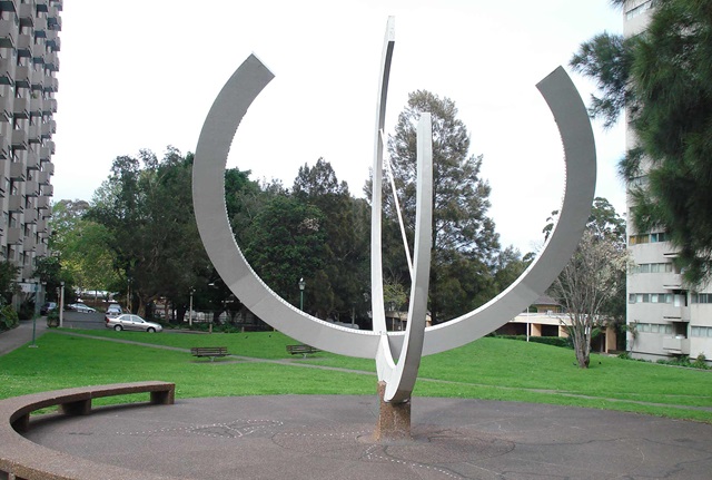 large cement and pebblecrete sundial made of hollow steel and painted white is at the centre of a circular pebblecrete area with seating around its perimeter.