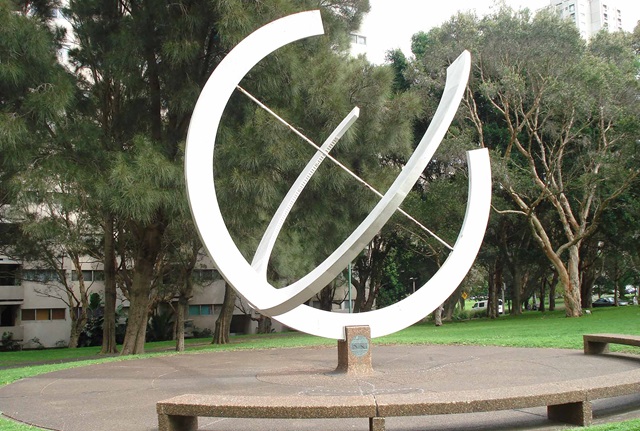 large cement and pebblecrete sundial made of hollow steel and painted white is at the centre of a circular pebblecrete area with seating around its perimeter.
