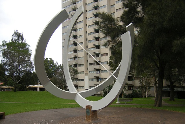 large cement and pebblecrete sundial made of hollow steel and painted white is at the centre of a circular pebblecrete area with seating around its perimeter.
