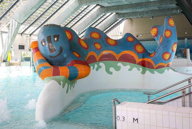 Large sculpture resembling a monster covered in blue red and orange tiles situated in an indoor swimming pool