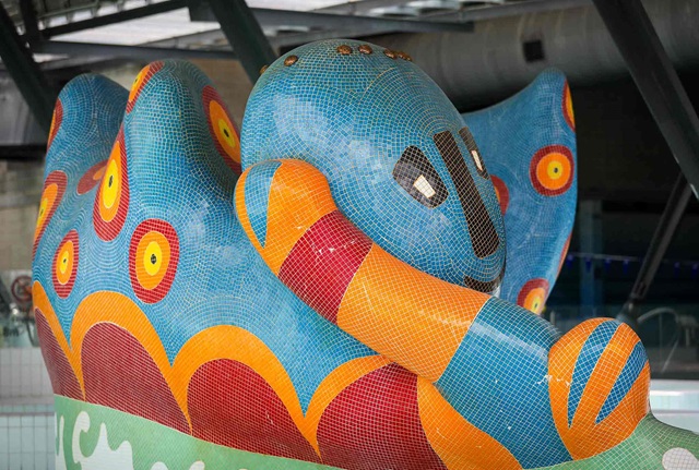 Large sculpture resembling a monster covered in blue red and orange tiles situated in an indoor swimming pool
