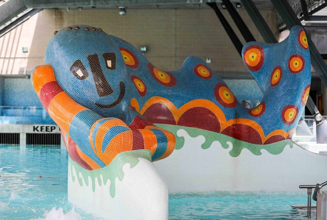 Large sculpture resembling a monster covered in blue red and orange tiles situated in an indoor swimming pool