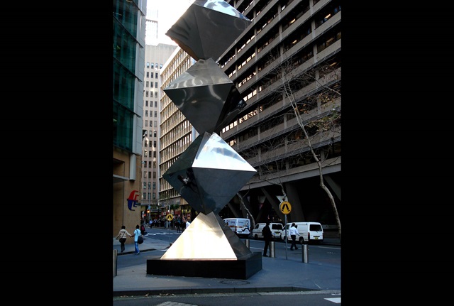 A tower sculpture made from stainless steel nearly 20 metres high sits at a busy city intersection between tall buildings