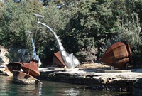 Two cone-shaped, rusted steel sculptures resembling giant fishing bobbers rest on the shore of a bay. Two more similar sculptures rest in the water, attached by cables to 2 stainless steel sculptures shaped like abstracted cranes.
