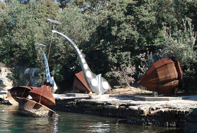 Two cone-shaped, rusted steel sculptures resembling giant fishing bobbers rest on the shore of a bay. Two more similar sculptures rest in the water, attached by cables to 2 stainless steel sculptures shaped like abstracted cranes.