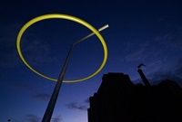 Sculpture of a tapered yellow ring balanced on a silver arm from a tall, tilted silver mast at night.