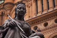 Close view of the upper half of a bronze sculpture of Queen Victoria, seated, wearing a crown and holding a sceptre and an orb. In the background is the sandstone facade of the Queen Victoria Building.