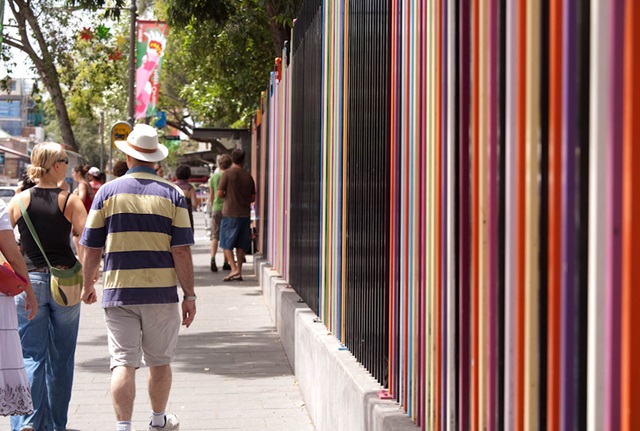 2 people walk on the footpath next to a highly coloured metal school fence with posts