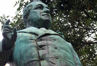 The upper half of a bronze statue of William Bede Dalley, viewed from below. Dalley is dressed in a great coat with one hand raised, and the statue has a strong patina from weathering.