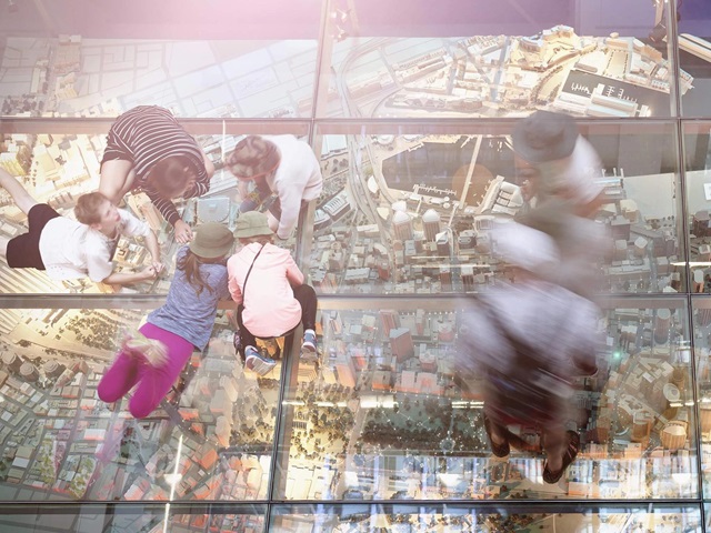 Two groups of people on the glass floor. They look at the city model displayed under the glass floor.  