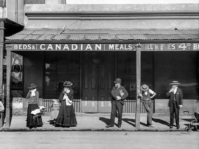People standing in a queue in front of The Canadian Dining Rooms on a sunny day.
