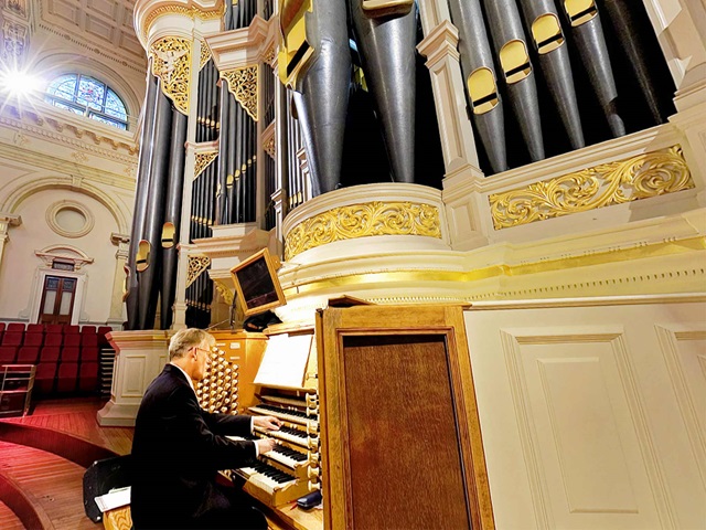 A seated man plays an organ, with pipes soaring above him.