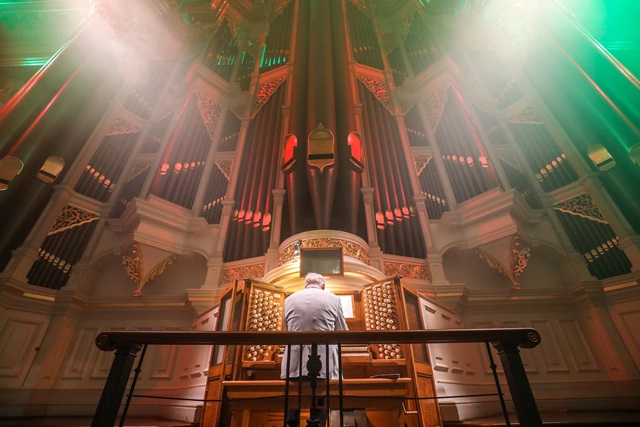 A man is seated at the keyboard of an organ surmounted by tall and ornate pipes.