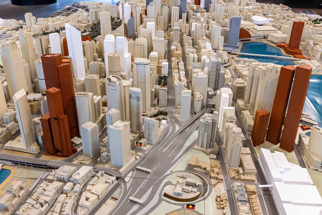 A detailed scale model of a modern city centre features high-rise buildings, roads, and waterways, with Australian Aboriginal flags visible on some structures.