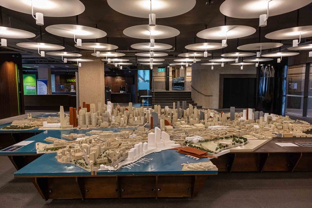A detailed architectural scale model of a cityscape with buildings, water features, and landscaped areas displayed indoors under modern ceiling lights.