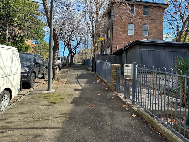 Narrow paved path beside parked cars and a brick building. Leafless trees line the walkway, and a metal fence encloses a small area on the right.