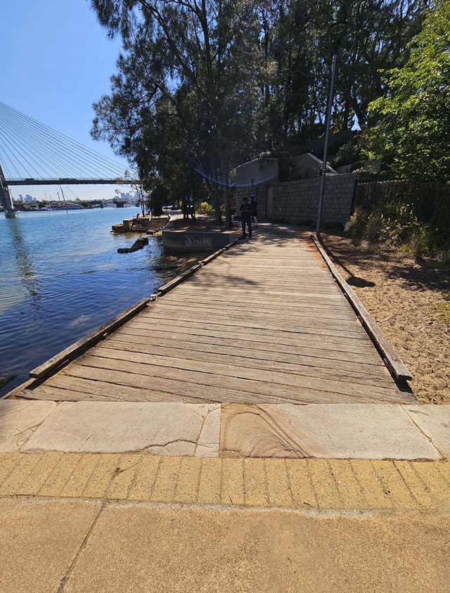 A wooden walkway extends over water, bordered by trees and a sandy area, with a bridge visible in the background under a clear sky.
