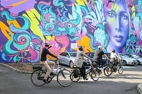 People riding bicycles past a wall of artwork.
