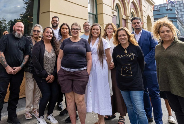A group of thirteen people stands together outside a building, posing and smiling for a group photo on a city street.