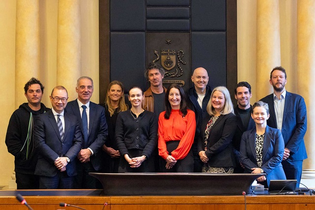 A group of twelve people in business attire pose for a photo in a formal meeting room with a crest displayed on the wall behind them.