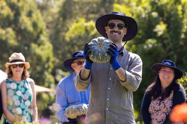 A man wearing gloves and a sunhat holds a pumpkin outdoors, smiling at the camera, with three other people in hats standing behind him.