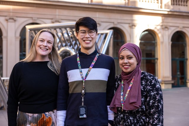 Three City of Sydney employees stand together on the terrace of Town Hall House, smiling and wearing matching colourful lanyards. Behind them is a large, abstract stainless steel sculpture.