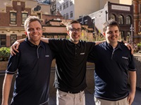 Three City of Sydney employees stand on a rooftop terrace at Town Hall House, with their arms around each other. They are wearing matching blue polo shirts and smiling.