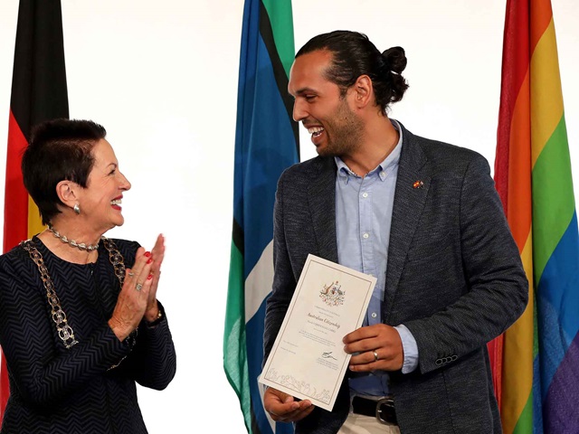 A smiling man holds a certificate, and a woman applauds him. They stand in front of flags.