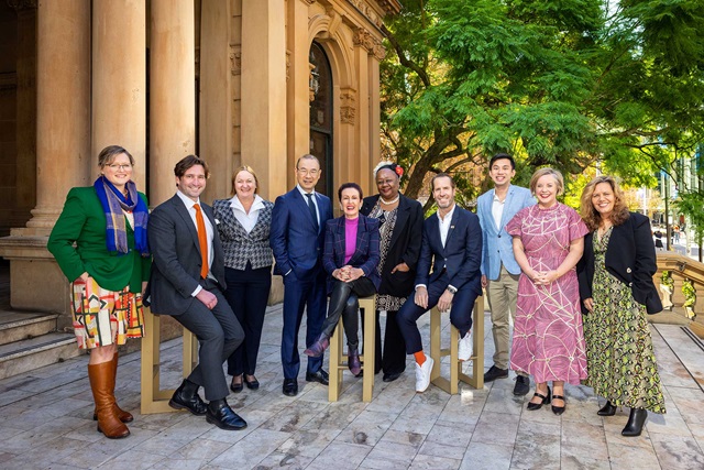 Lord Mayor and councillors stand in front of a heritage columned Sydney Town Hall building. Tree in the background.