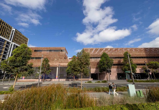 A modern building with a patterned façade stands behind trees and a road; two people walk on the pavement in the foreground under a partly cloudy sky.