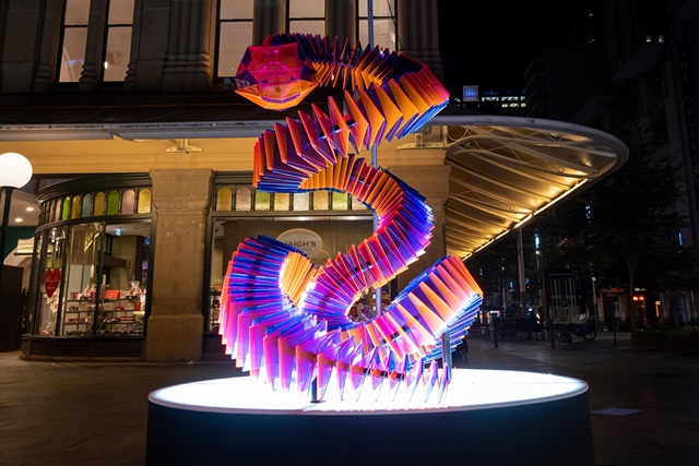 A large, colourful lantern shaped like an origami snake sits on a plinth outside the Queen Victoria Building at night. It is lit from below by a bright white light, and its orange, violet, pink and blue translucent panels catch the light and seem to glow.