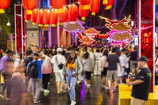 A woman stands smiling in a busy, lantern-lit street decorated with neon dragon signs during a night time festival, surrounded by blurred crowds.