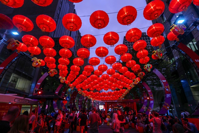 Crowds gather under numerous red lanterns hanging above a city street at dusk for a festival. Tall buildings and decorative arches are visible in the background.