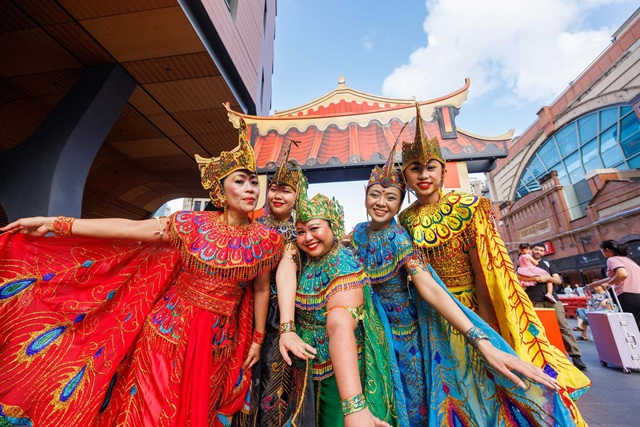 Five women in colourful, ornate traditional costumes pose together outdoors in front of a decorative structure and modern buildings.