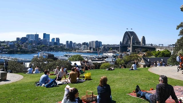 Groups of people sit on the grass with picnics, overlooking Sydney Harbour with views to the North