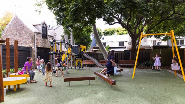 Children and adults at a playground with swings, slides, and benches, surrounded by trees.
