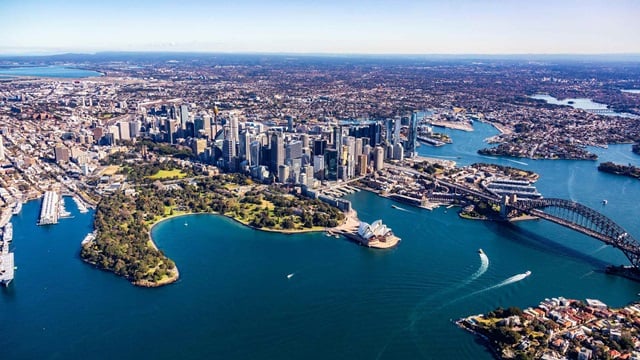 Aerial view of Sydney showing the central business district, Sydney Opera House, Harbour Bridge, and surrounding waterways and urban areas under a clear sky.