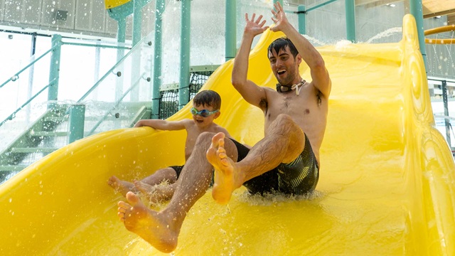 Parent and child play on a yellow water slide at a swimming pool